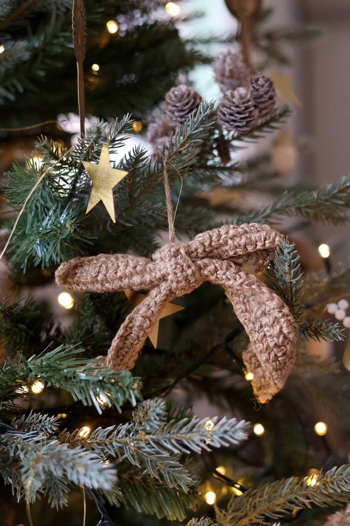 A handmade brown crochet ornament shaped like a bow with small gold star accents, hanging on a decorated Christmas tree with lights and pinecones.
