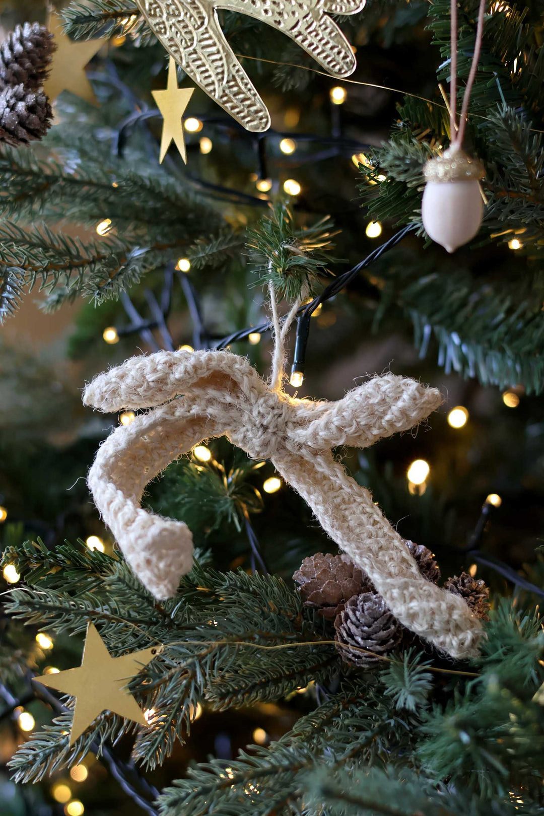 A beige knitted Christmas ornament shaped like a bow or ribbon, hanging on a decorated evergreen tree with warm string lights, pinecones, and gold star ornaments.