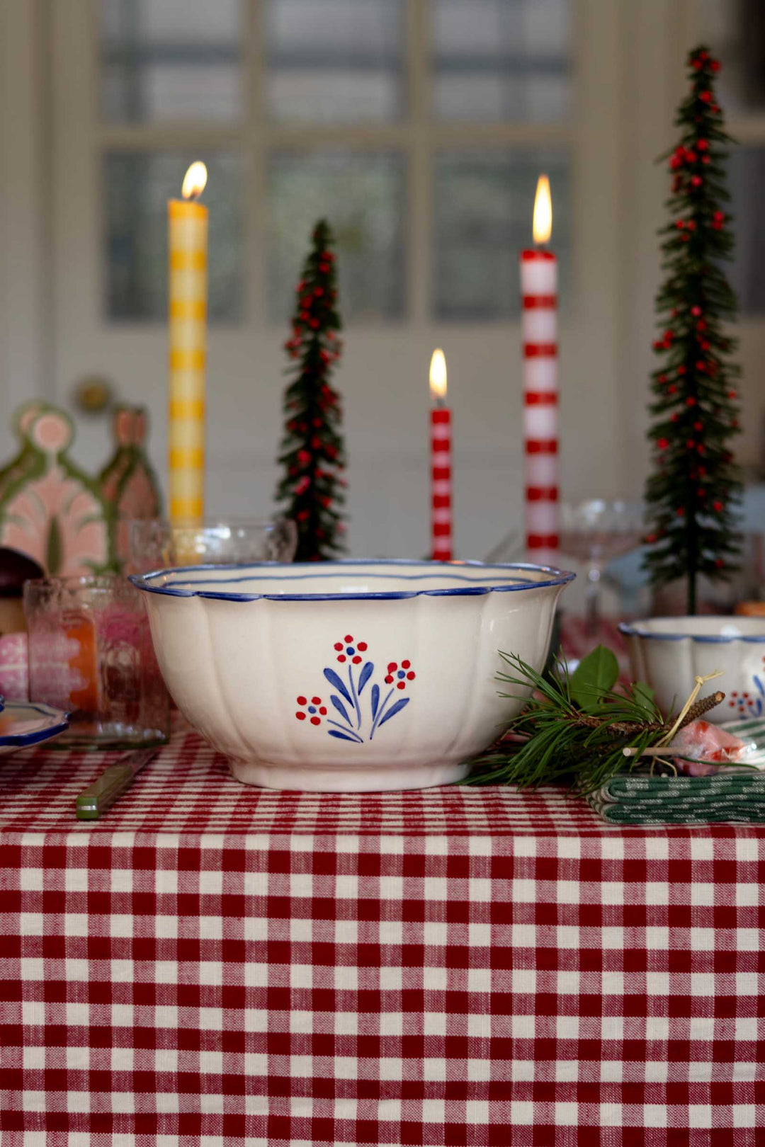 A white ceramic bowl with a blue rim and a red and blue floral pattern, set on a red and white checkered tablecloth with holiday decorations including candles and miniature Christmas trees.