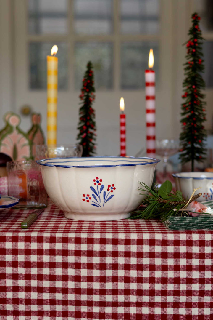 A white ceramic bowl with a blue rim and a red and blue floral pattern, set on a red and white checkered tablecloth with holiday decorations including candles and miniature Christmas trees.