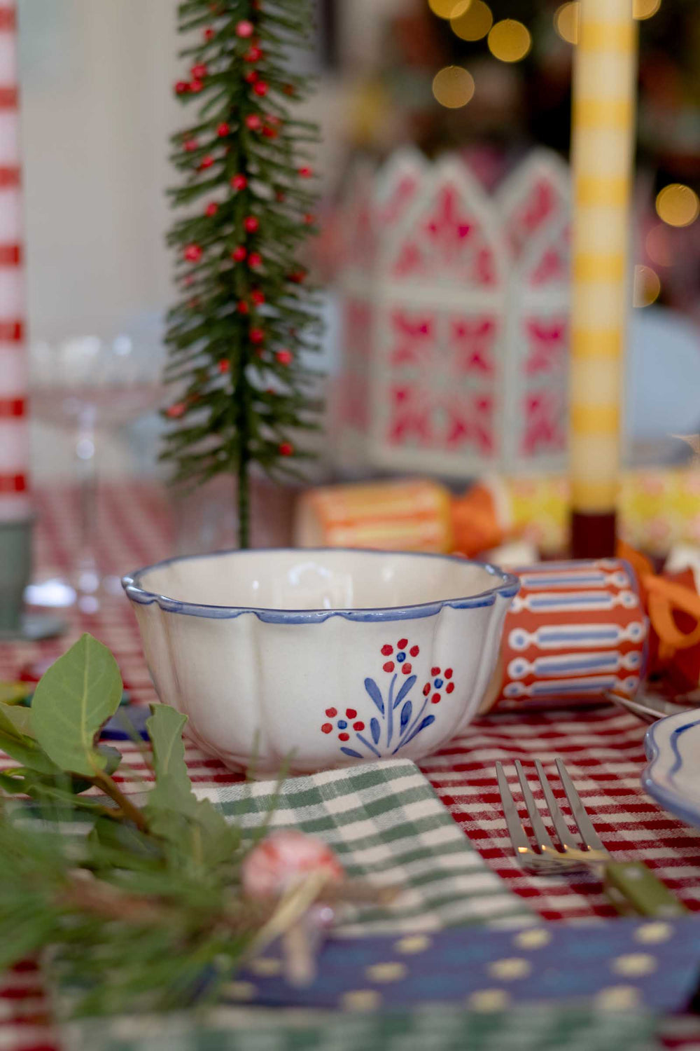 A white ceramic bowl with a blue scalloped rim and red floral pattern, set on a red and white checkered tablecloth with a green gingham napkin and a small Christmas tree in the background.