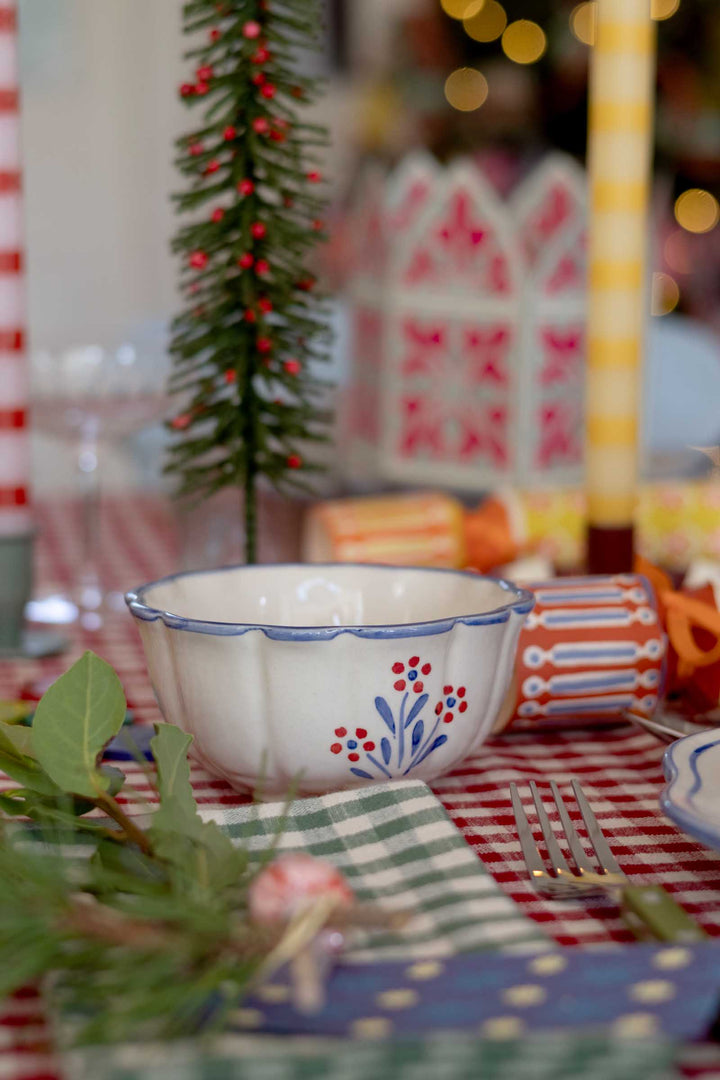A white ceramic bowl with a blue scalloped rim and red floral pattern, set on a red and white checkered tablecloth with a green gingham napkin and a small Christmas tree in the background.