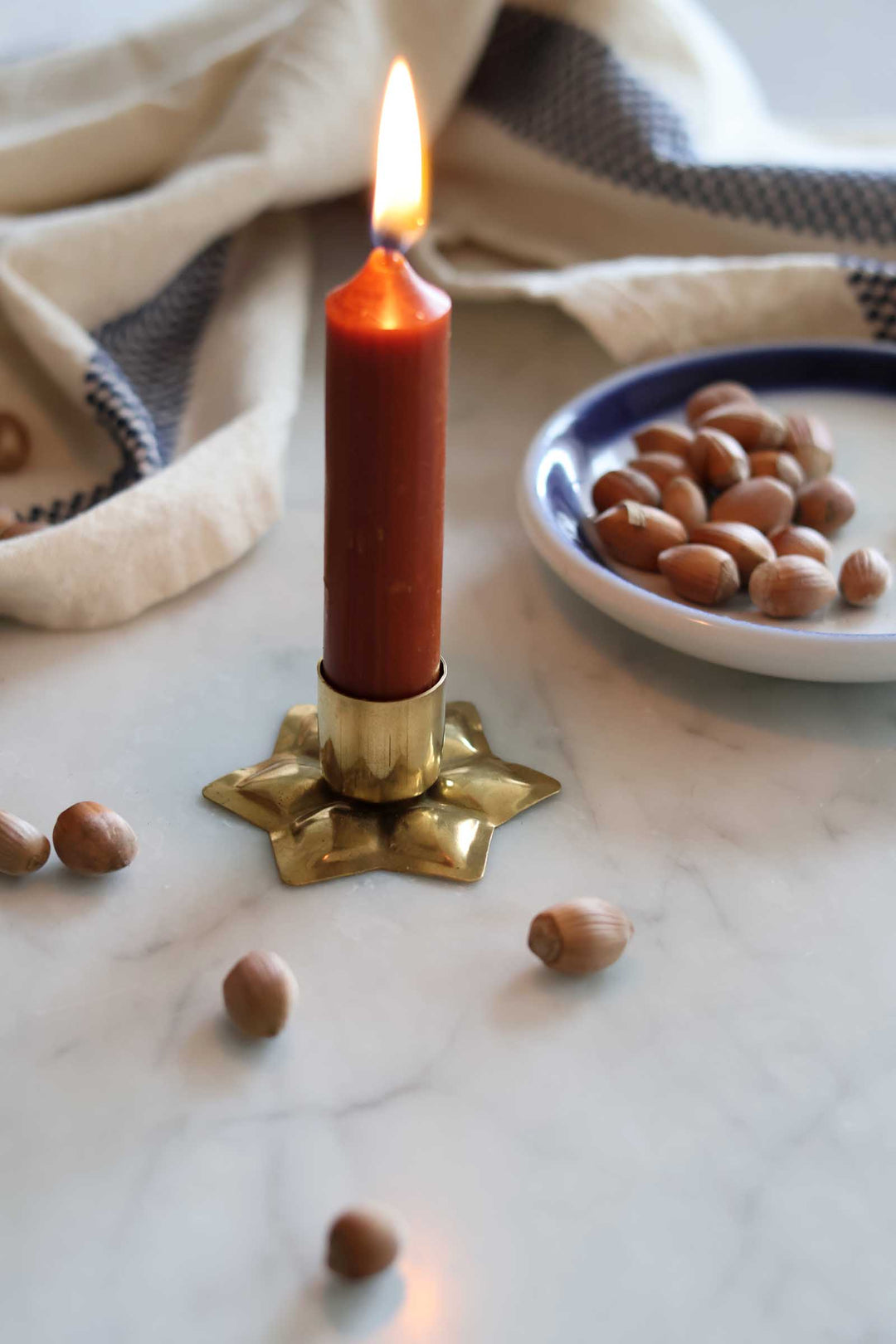 A lit brown candle in a gold star-shaped holder on a marble surface, with scattered hazelnuts and a small plate of hazelnuts nearby, and a cream cloth in the background.