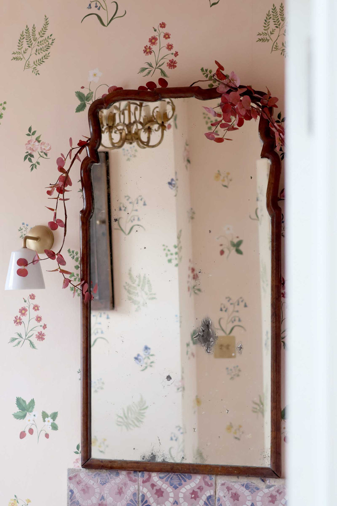 A vintage-style rectangular wall mirror with a dark brown wooden frame and decorative floral garlands, mounted on a pink floral wallpapered wall next to a gold sconce.
