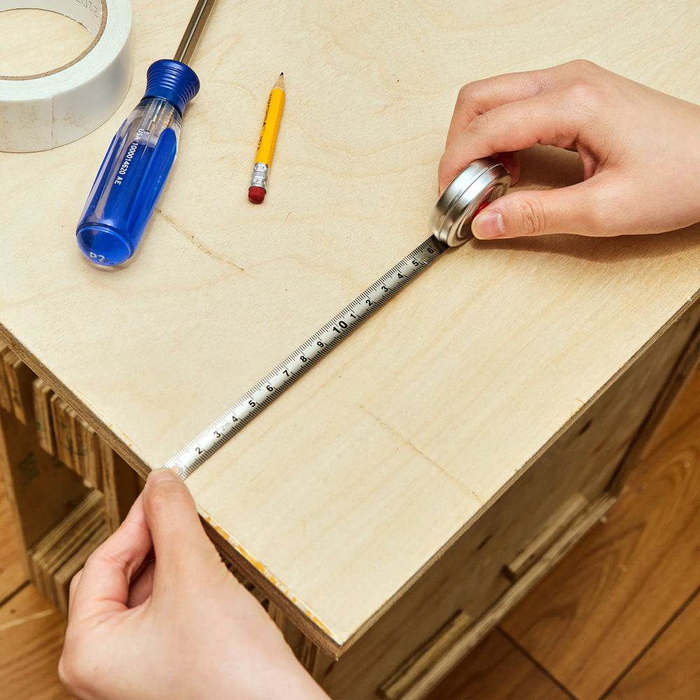 Person measuring a wooden box with a tape measure, using tools like a screwdriver and pencil on a wooden surface.