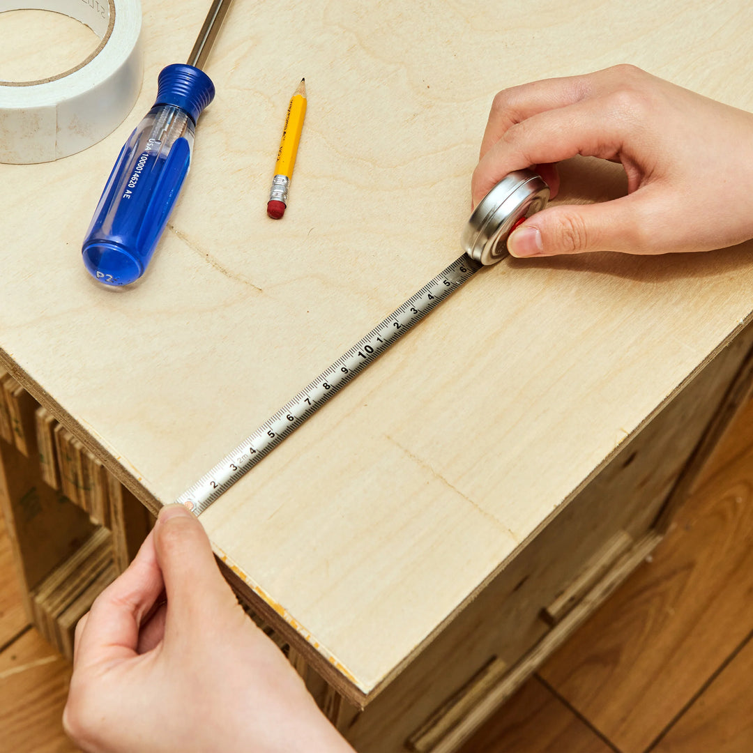 Person measuring a wooden box with a tape measure, using tools like a screwdriver and pencil on a wooden surface.