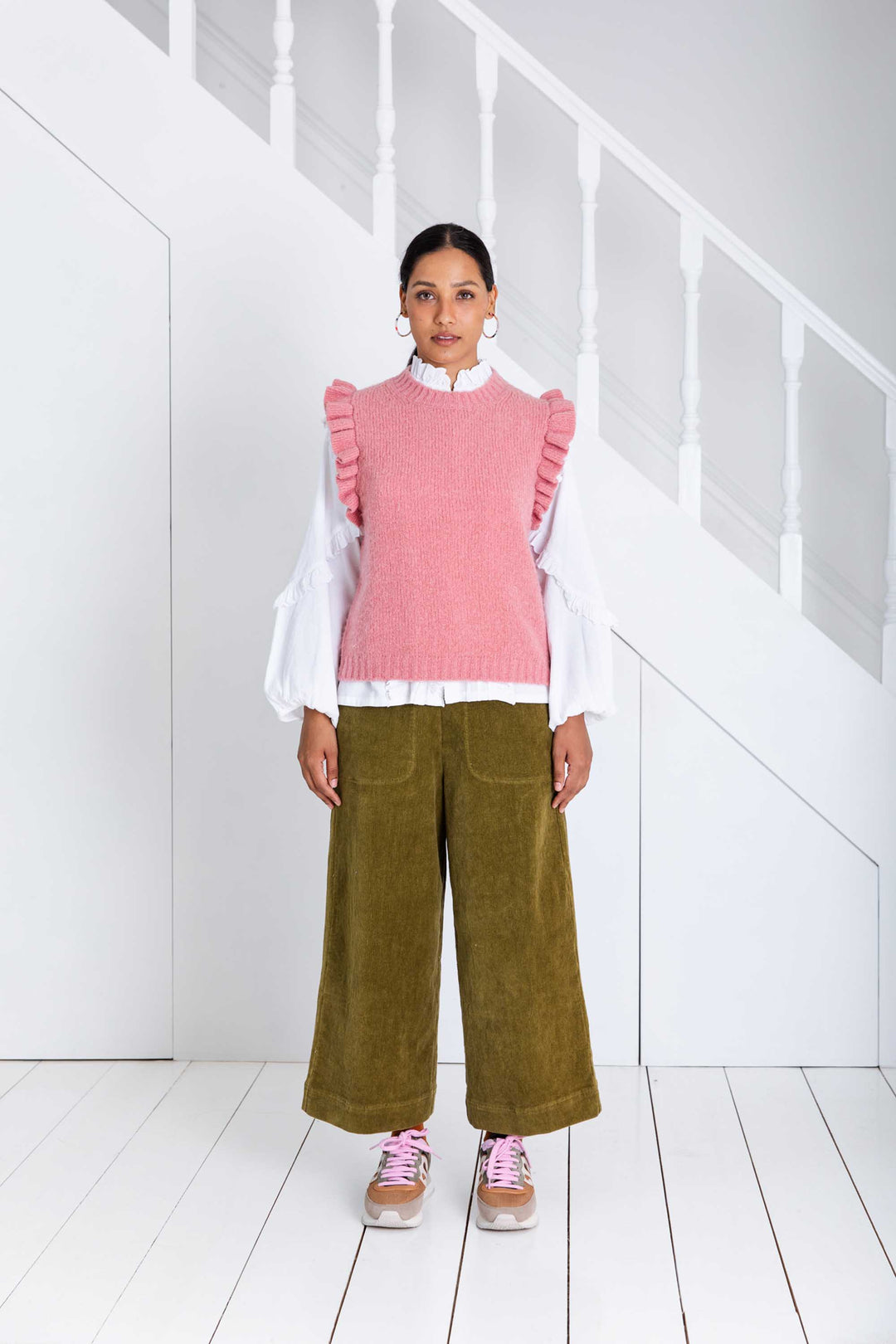 Person wearing a pink ruffled vest over a white shirt with green pants in a minimalistic setting.