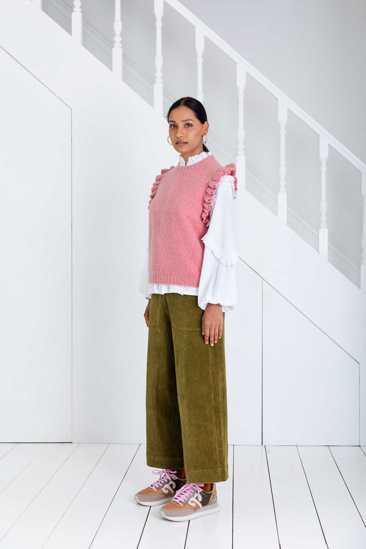 Person wearing a pink sweater with ruffled details, white shirt, green pants, and brown shoes in a minimalistic setting.