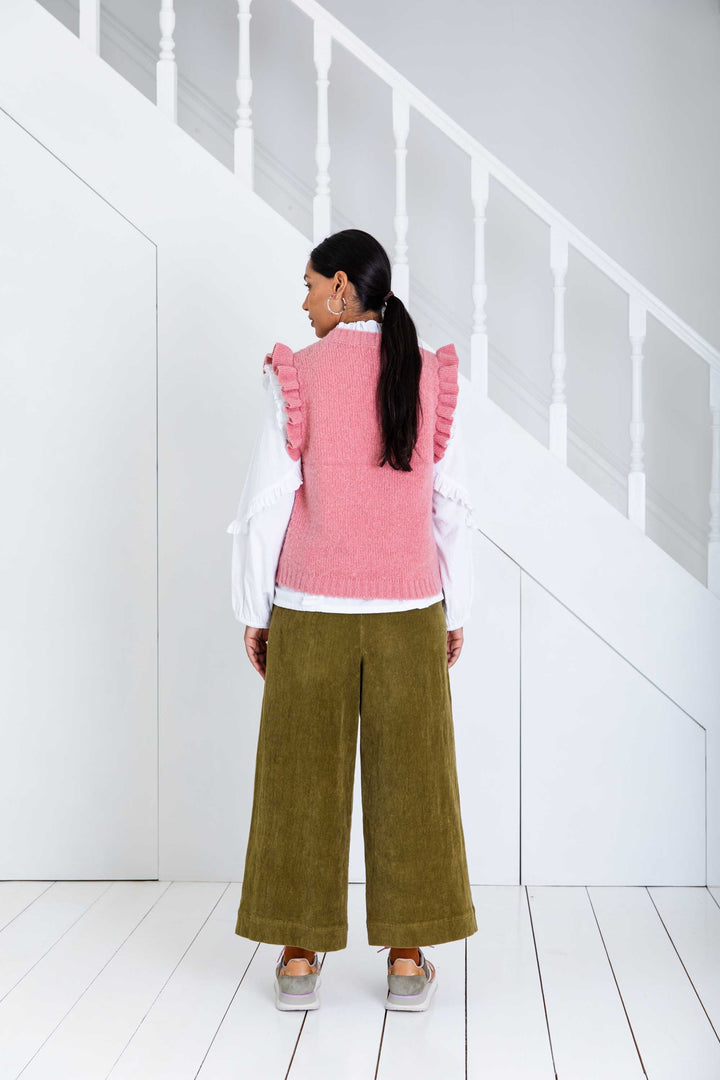 Person wearing a pink sweater and green pants standing in a white room.