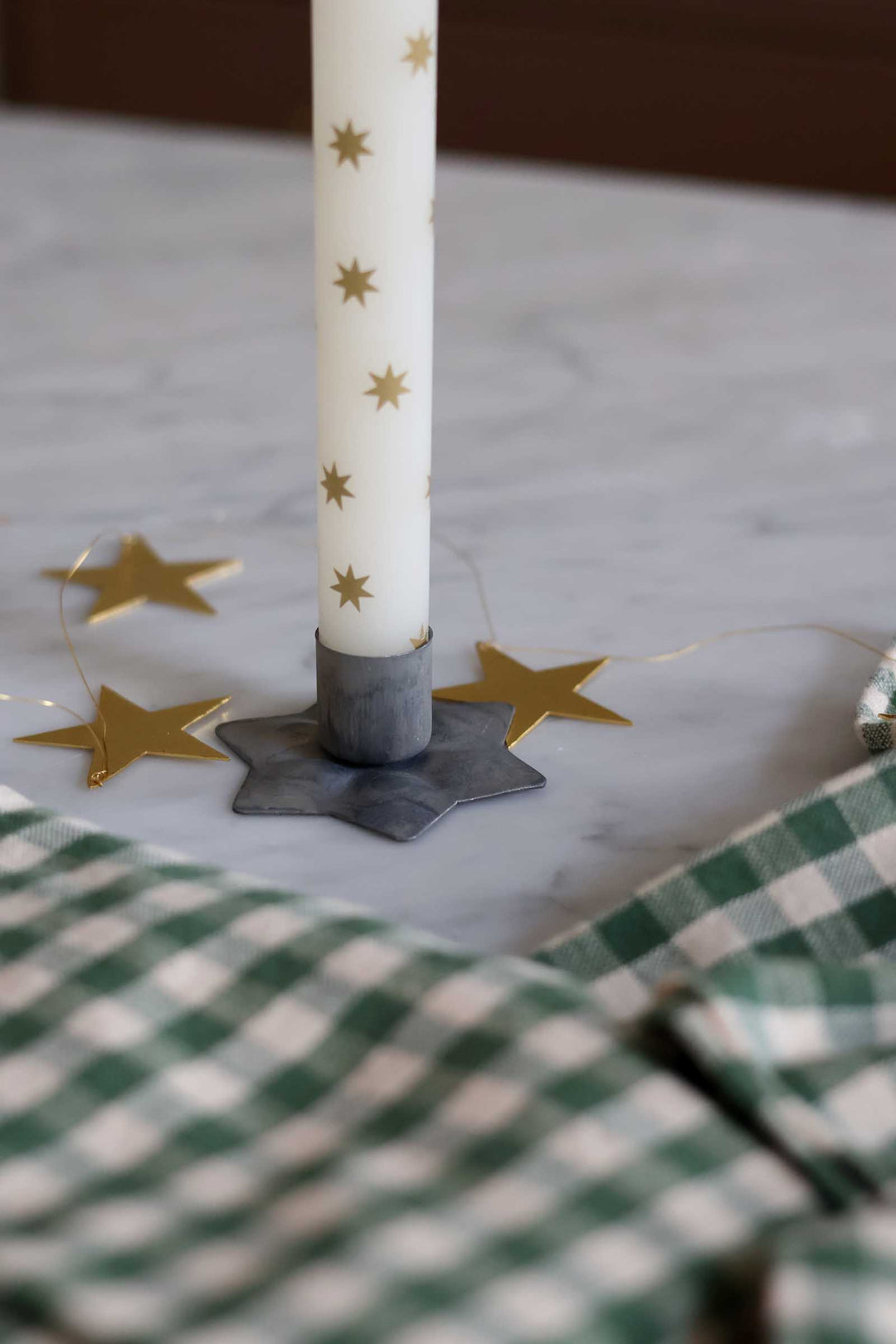 A white taper candle with small gold star motifs stands in a dark gray star-shaped metal holder on a light marble surface. Thin gold star garlands are draped nearby, and a green-and-white checkered cloth is visible in the foreground.