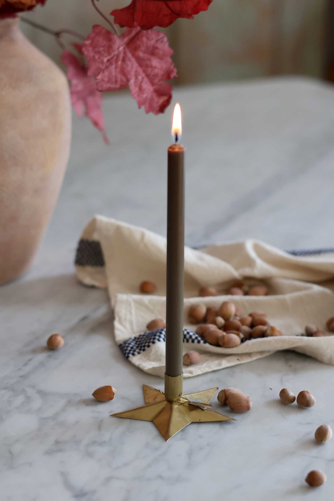 A single lit taper candle in a dark brown glass holder stands on a gold star-shaped base on a marble surface, styled with hazelnuts, a beige cloth, and autumn leaves in the background.