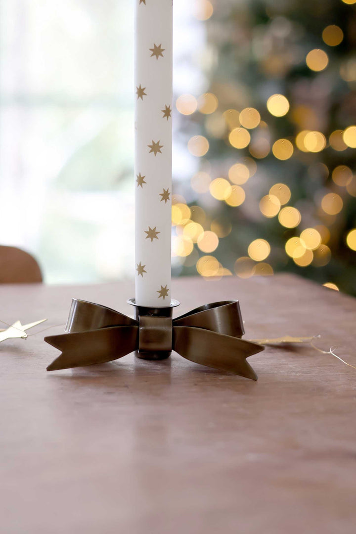 A white taper candle decorated with small gold stars, set in a dark metallic holder with a matching ribbon bow, placed on a wooden table with a blurred Christmas tree and warm lights in the background.