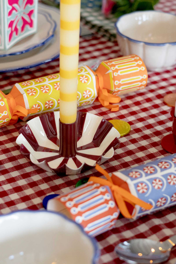 A close-up of a decorated table setting with a red-and-white checkered tablecloth. A striped candle in a scalloped white, brown, and red holder is centered, surrounded by colorful patterned cylindrical ornaments tied with orange ribbons and white bowls.