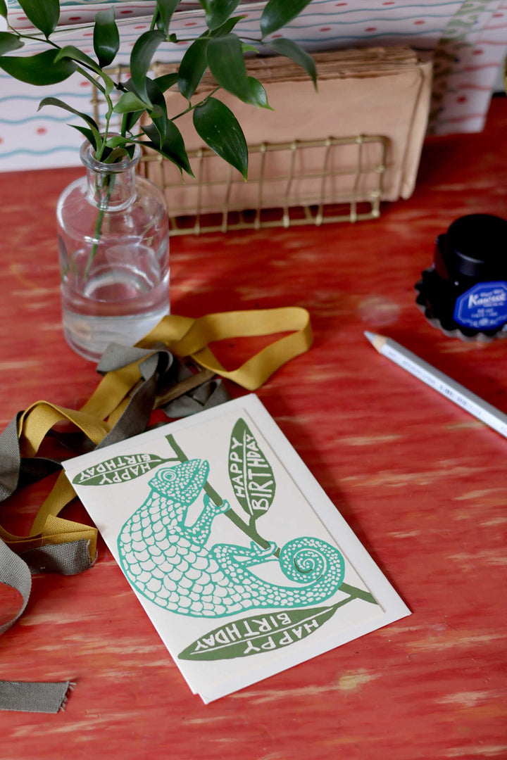 Card with green lizard design on a red surface with a plant and bottle in the background