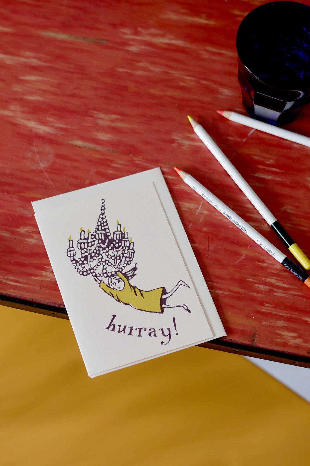 Note card with illustration and 'hurray!' text on a red surface with pens.