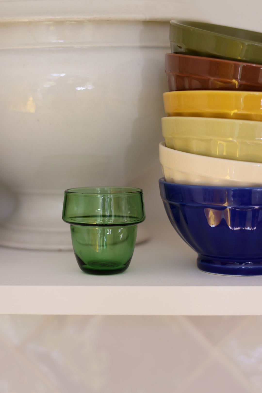 A small translucent green glass tumbler sits on a white shelf next to a stacked set of colorful bowls in olive green, brown, yellow, cream, and dark blue.