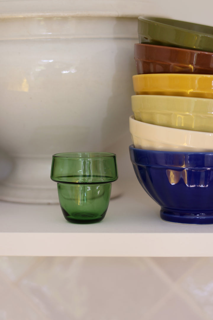 A small translucent green glass tumbler sits on a white shelf next to a stacked set of colorful bowls in olive green, brown, yellow, cream, and dark blue.