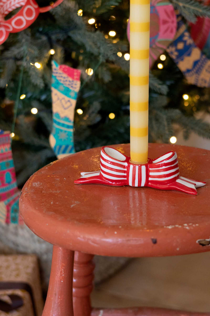 A close-up of a red and white striped ceramic candle holder shaped like a bow, holding a yellow striped candle on a rustic reddish-brown wooden stool, with a blurred Christmas tree in the background.