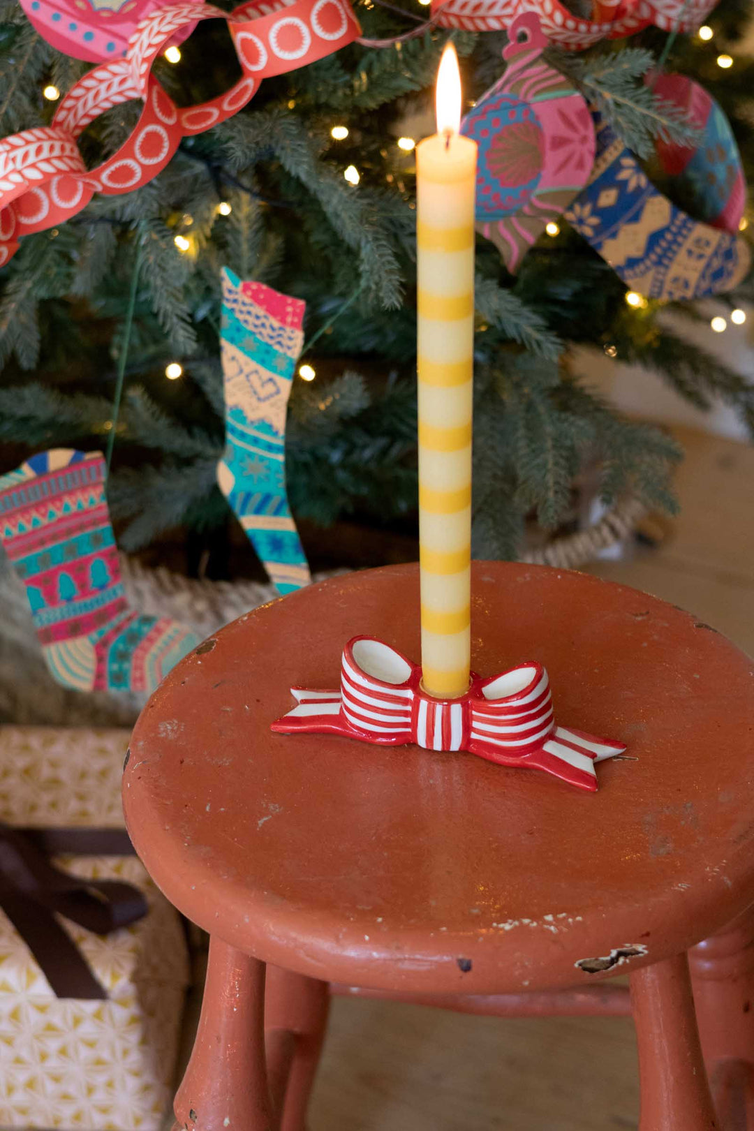 A lit yellow-and-white striped taper candle in a small red-and-white bow holder, placed on a rustic reddish-brown stool in front of a decorated Christmas tree.