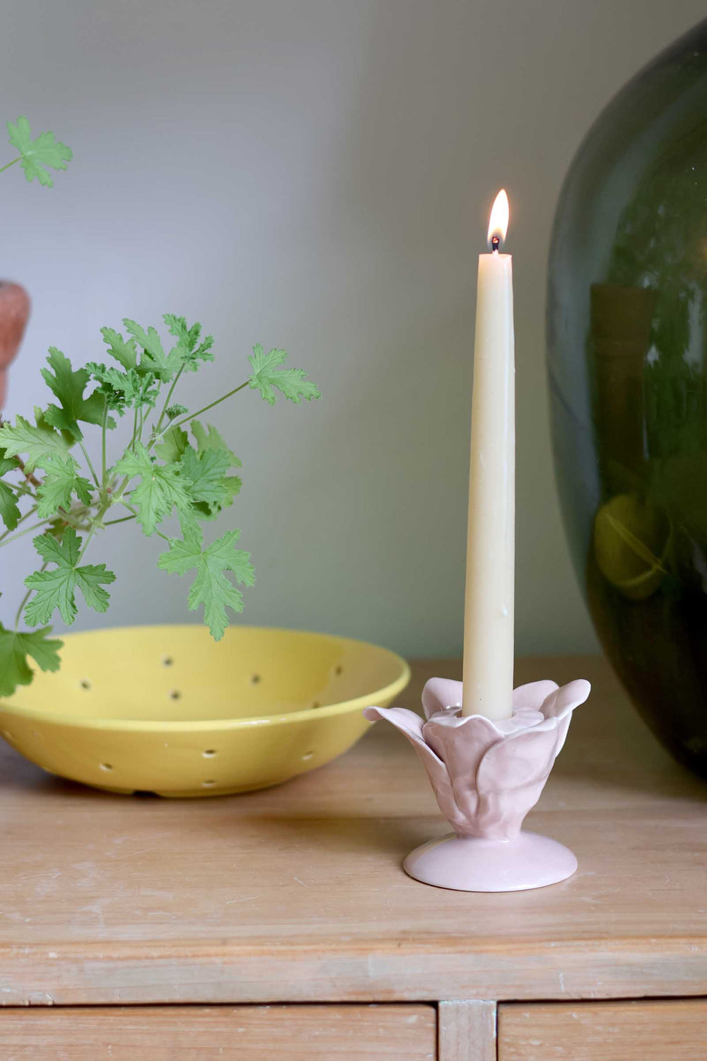 A lit cream-colored taper candle in a pale pink lotus-shaped holder, styled on a wooden table next to a yellow bowl and green leaves.
