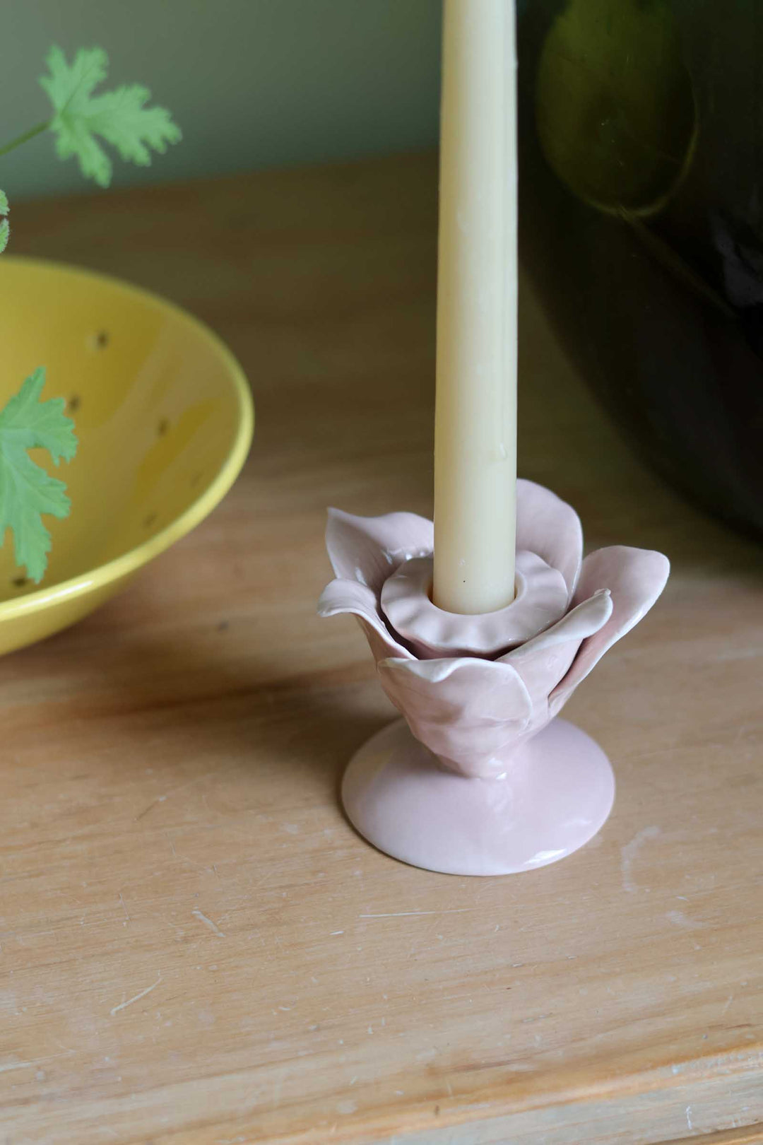 A pale pink ceramic candle holder shaped like a flower, holding a cream-colored candle, set on a light wooden tabletop with a yellow bowl in the background.