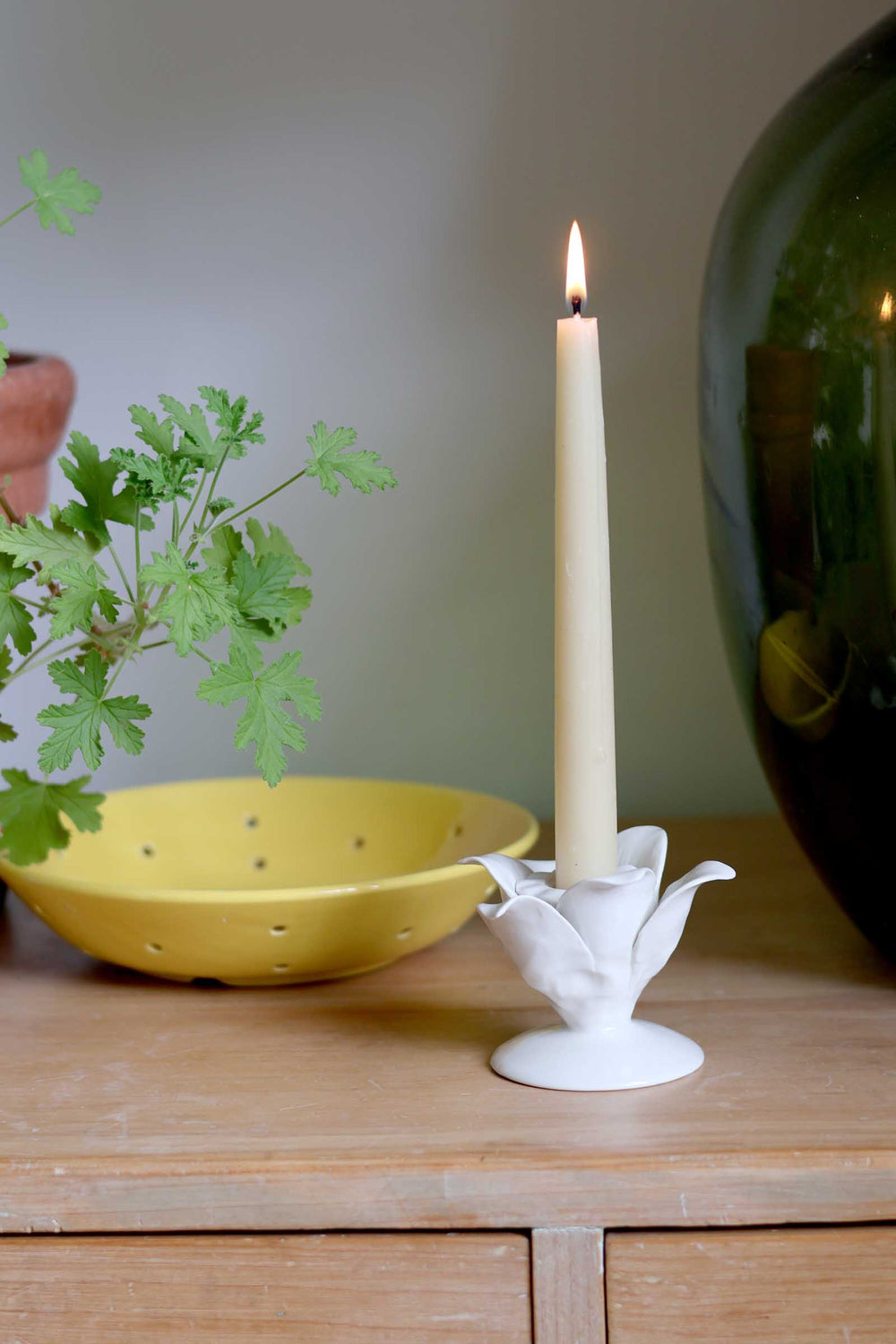 A lit cream taper candle in a white sculptural flower-shaped holder, styled on a wooden surface next to a yellow bowl and green leaves.