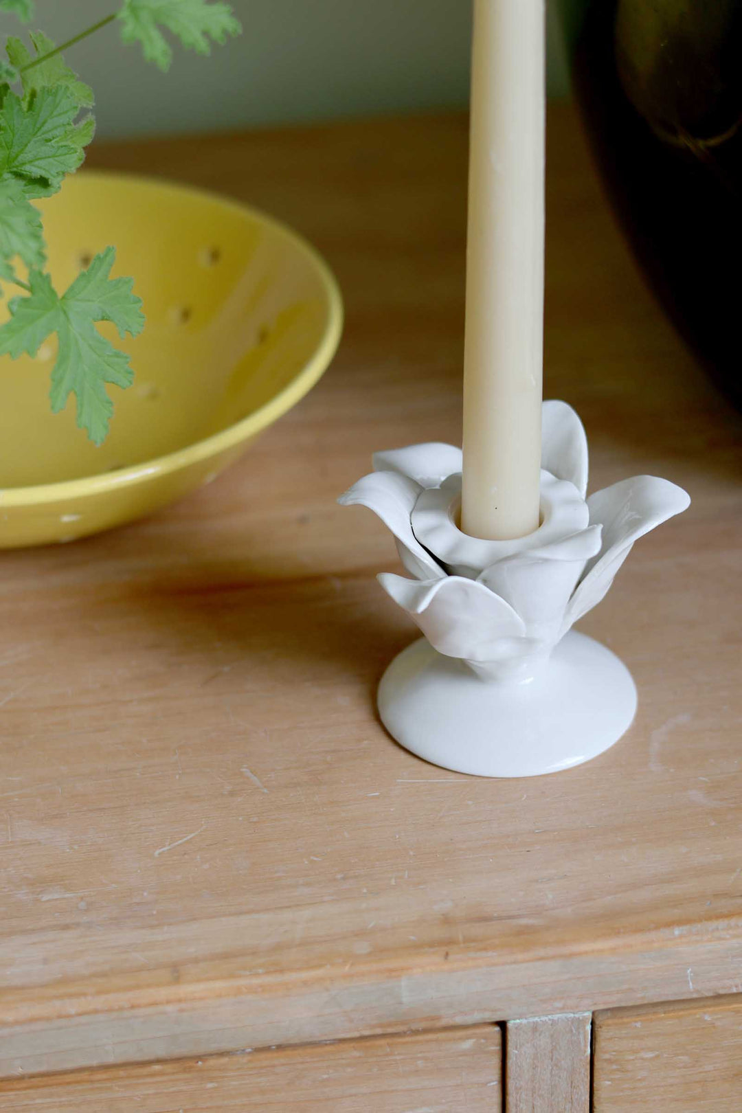 A white ceramic candle holder shaped like a lotus flower holding a cream-colored candle, set on a light wooden table next to a yellow bowl.