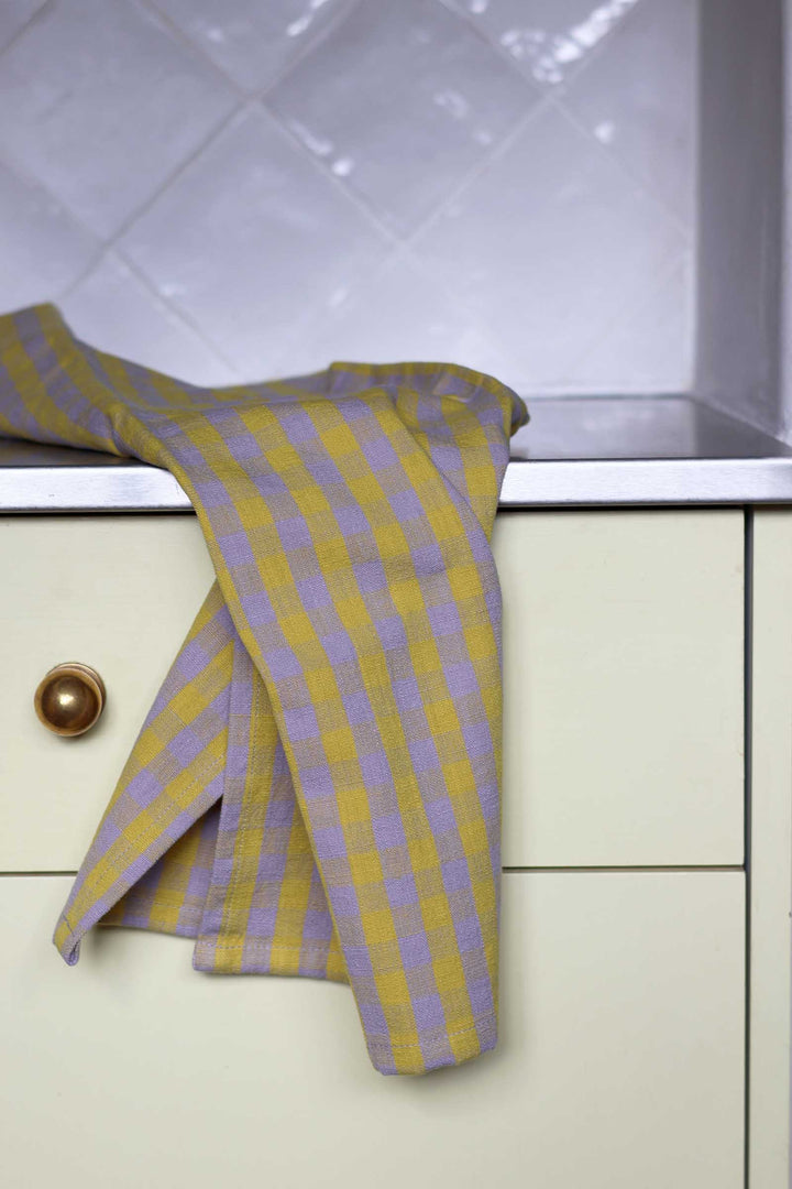 A yellow and lavender checkered kitchen towel draped over a cream-colored cabinet drawer with a gold knob, set against a white tiled backsplash.