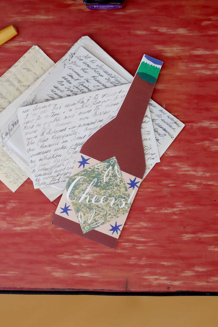 Decorative paper cutout of a bottle with 'Cheers' text on a red surface