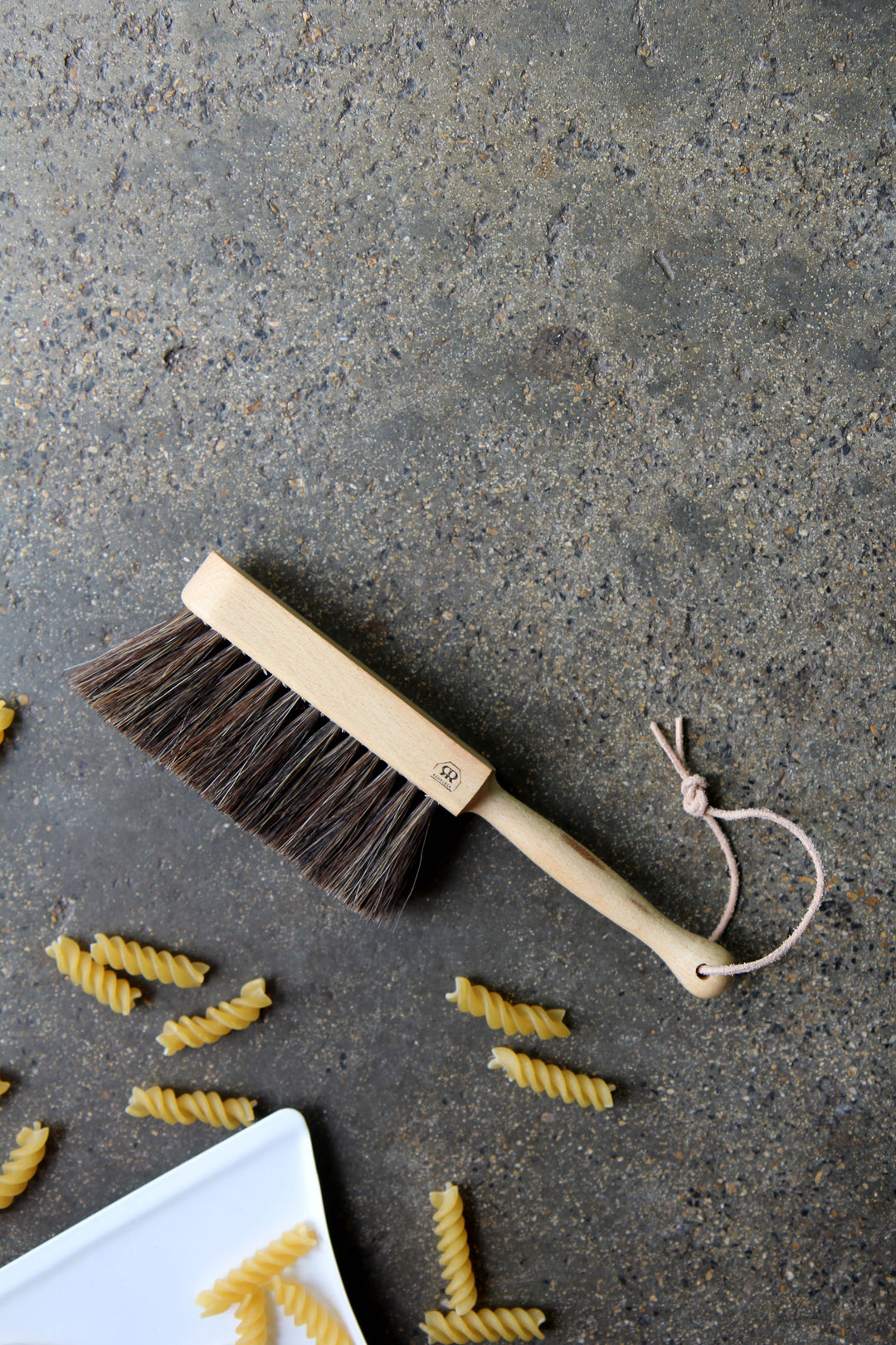 Dustpan with horsehair bristles on a concrete surface with pasta pieces.