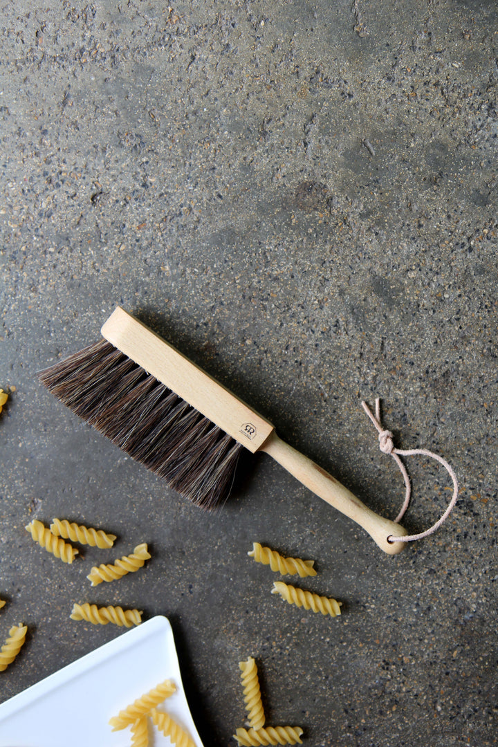 Dustpan with horsehair bristles on a concrete surface with pasta pieces.