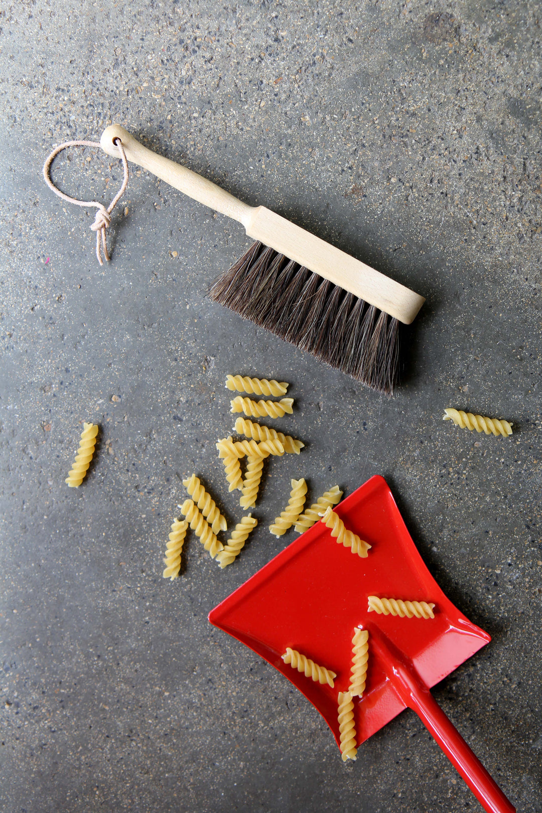 Cleaning tools including a brush and dustpan on a gray surface with scattered pasta.