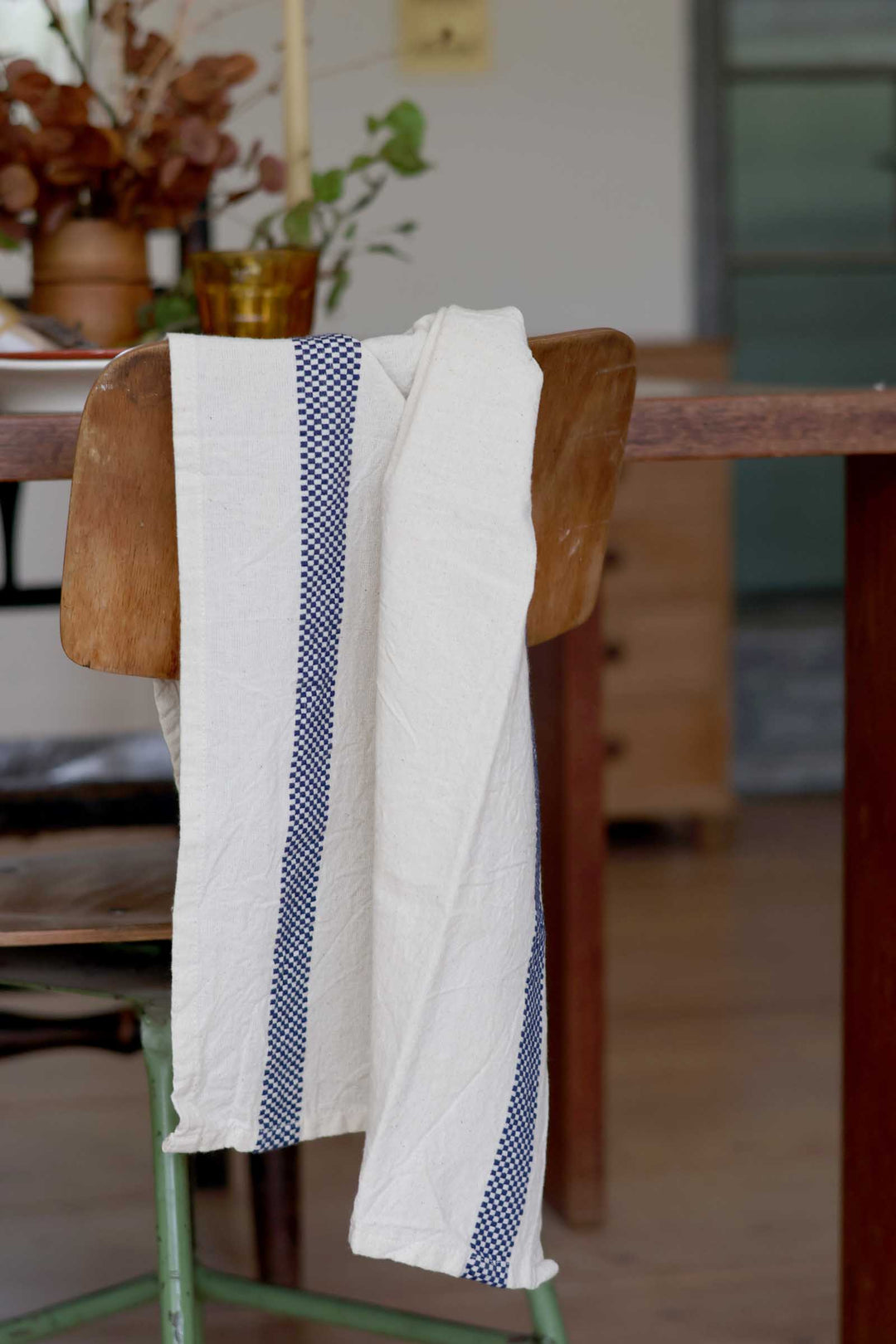 A white cotton tea towel with blue checkered stripes hangs over a rustic wooden chair in a softly blurred indoor setting.