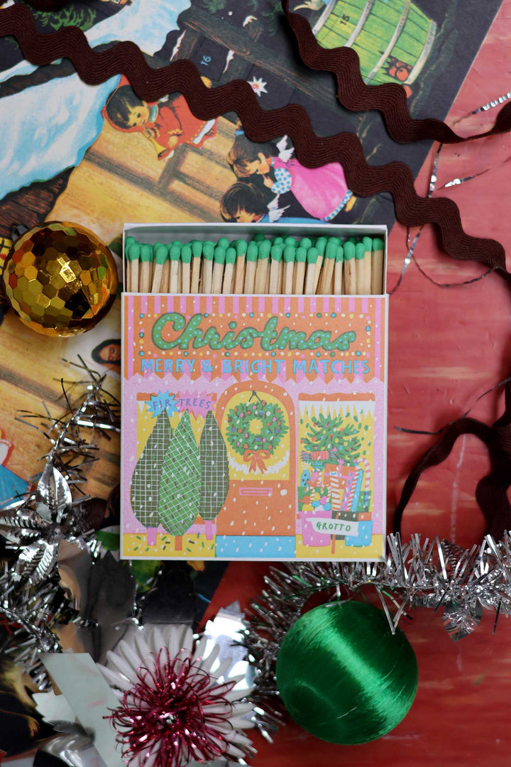 A Christmas-themed matchbox with green-tipped matches, surrounded by festive tinsel, ornaments, and holiday decorations on a red surface.
