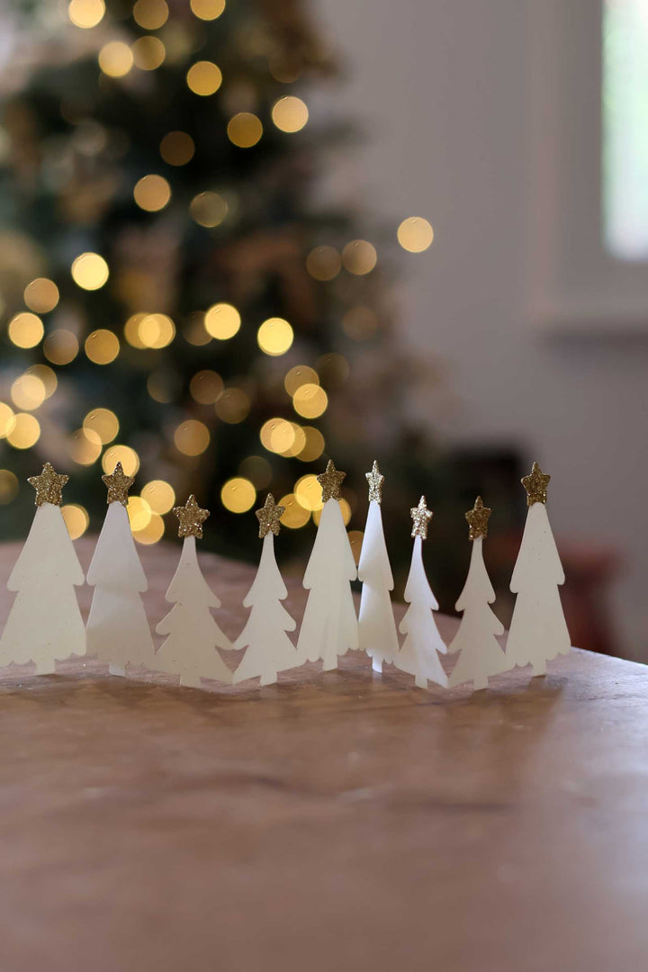 A row of small white paper Christmas tree ornaments with gold star tops, arranged on a wooden surface with a blurred Christmas tree and warm bokeh lights in the background.