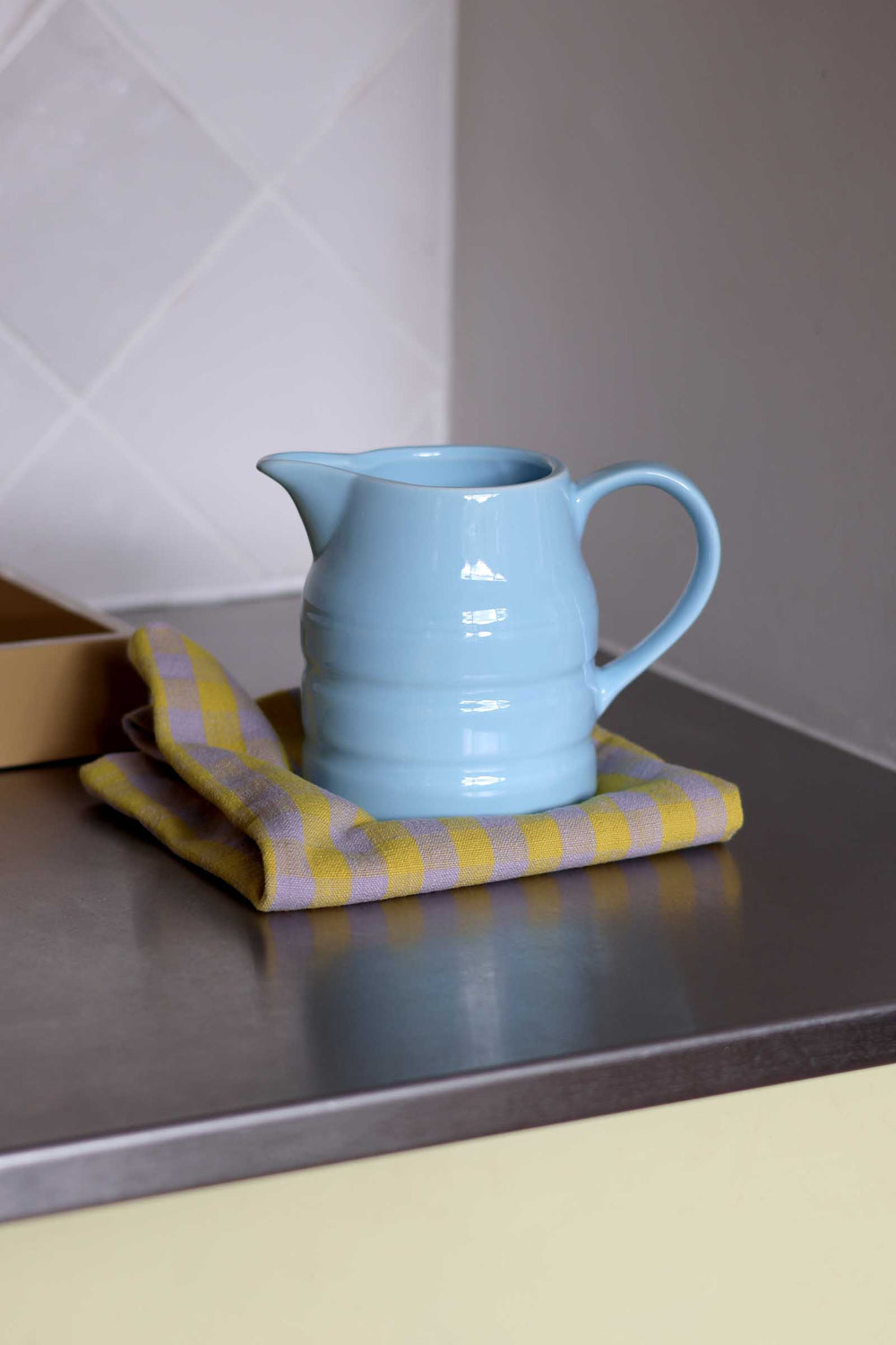 A light blue ceramic pitcher with a ribbed texture and curved handle, resting on a yellow and white striped cloth on a dark countertop.
