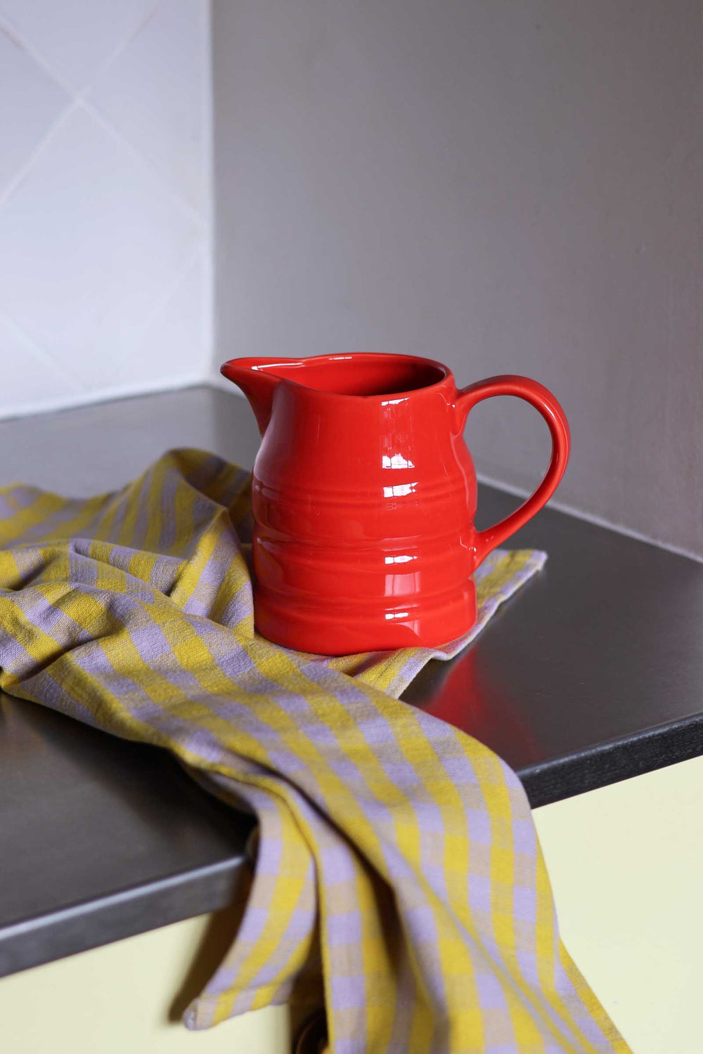 A glossy red ceramic pitcher with a curved handle sits on a dark countertop next to a yellow and white checkered cloth.