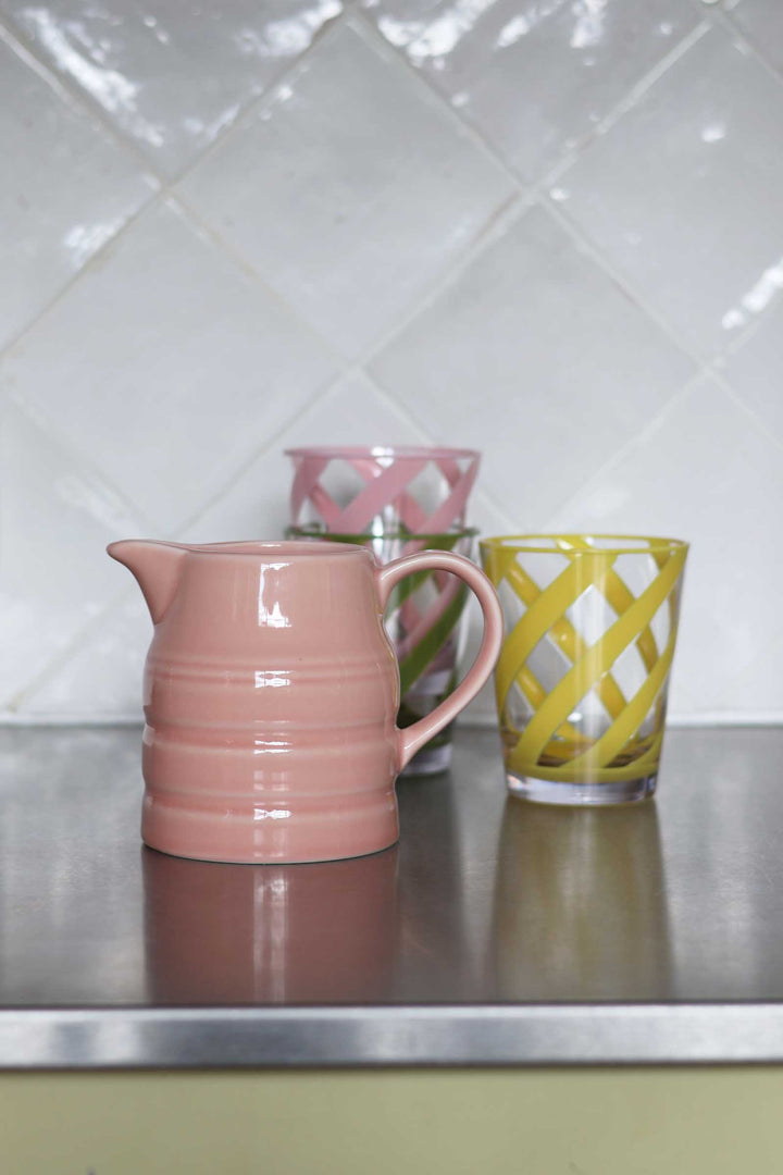 A pink ceramic pitcher with horizontal ridges stands on a metallic countertop next to a yellow lattice-patterned glass, with two more patterned glasses blurred in the background against a white tiled wall.