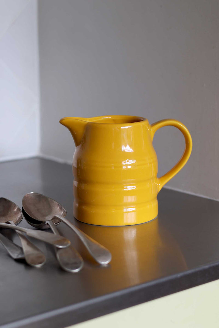 A glossy mustard yellow ceramic pitcher with a ribbed design, set on a dark gray countertop next to four silver spoons against a neutral gray background.