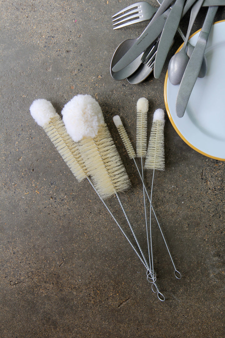Set of dish brushes on a concrete surface with cutlery and plates in the background.