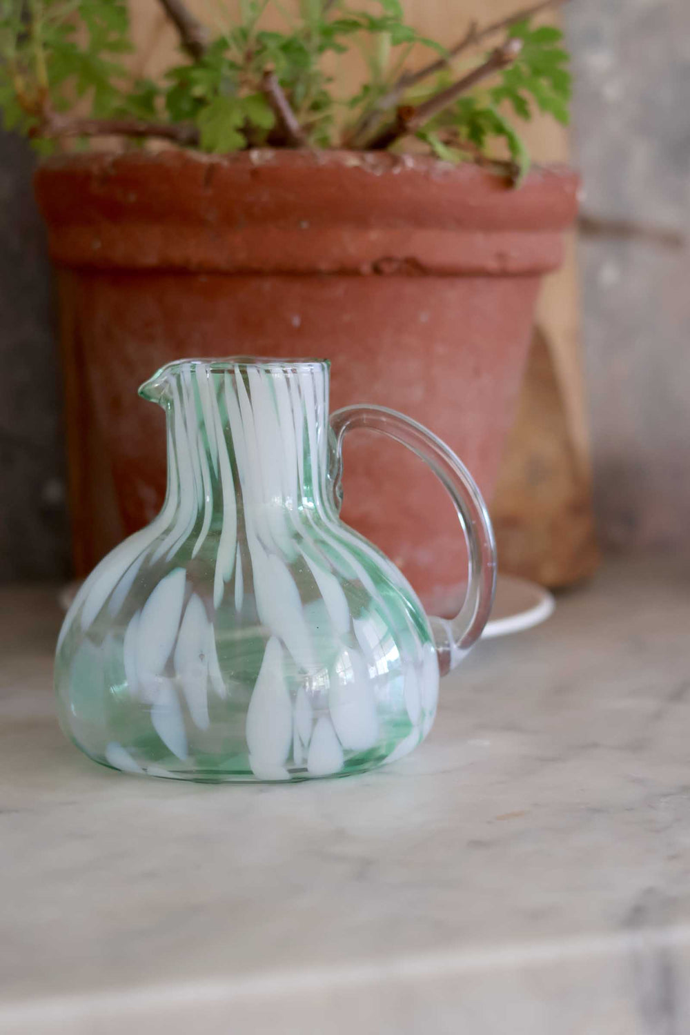 Clear glass pitcher with white floral design on a marble surface, potted plant in the background.