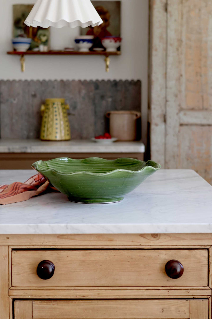 A green ceramic bowl with a scalloped rim sits on a white marble countertop in a rustic kitchen setting with a light wood cabinet below.