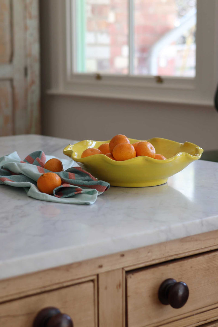 A bright yellow ceramic bowl filled with oranges on a white marble countertop, next to a folded patterned cloth napkin with another orange, set in a kitchen environment with wooden drawers and a window in the background.