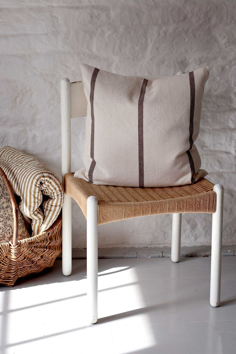 A beige linen throw pillow with two vertical dark brown stripes rests on a white-framed chair with a woven seat, set against a white textured brick wall.