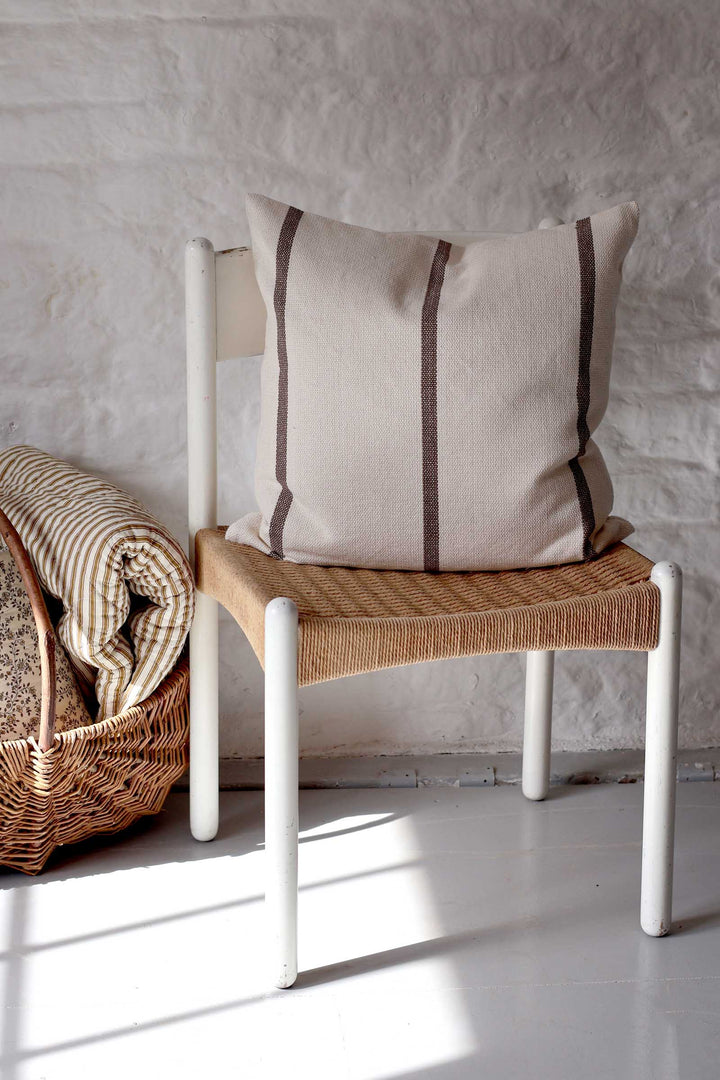 A beige linen throw pillow with two vertical dark brown stripes rests on a white-framed chair with a woven seat, set against a white textured brick wall.