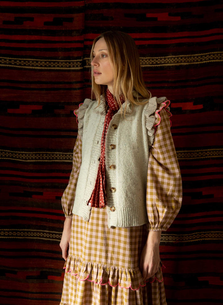 Woman wearing a checkered dress with a ruffled knitted gilet against a patterned wall.