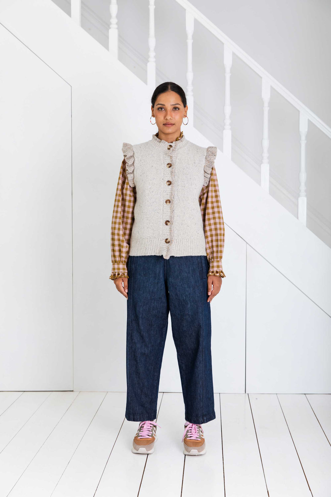Person wearing a plaid shirt, beige knitted vest, and blue jeans in a minimal white room.