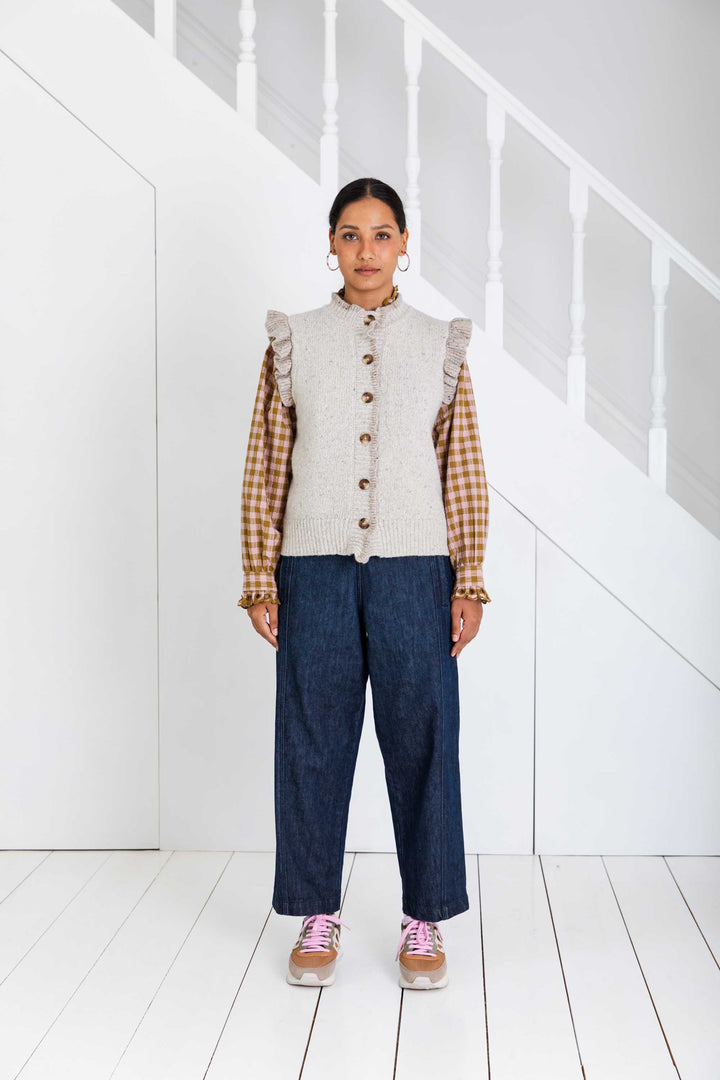 Person wearing a plaid shirt, beige knitted vest, and blue jeans in a minimal white room.