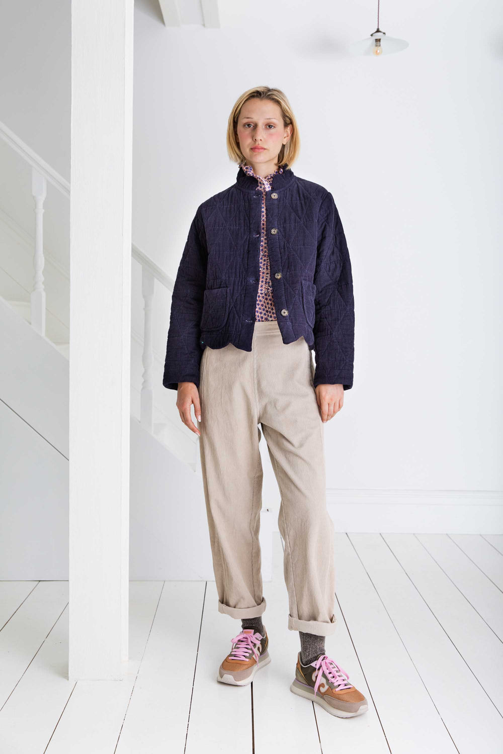 Person wearing a dark jacket, patterned shirt, beige pants, and pink sneakers in a minimal white room.