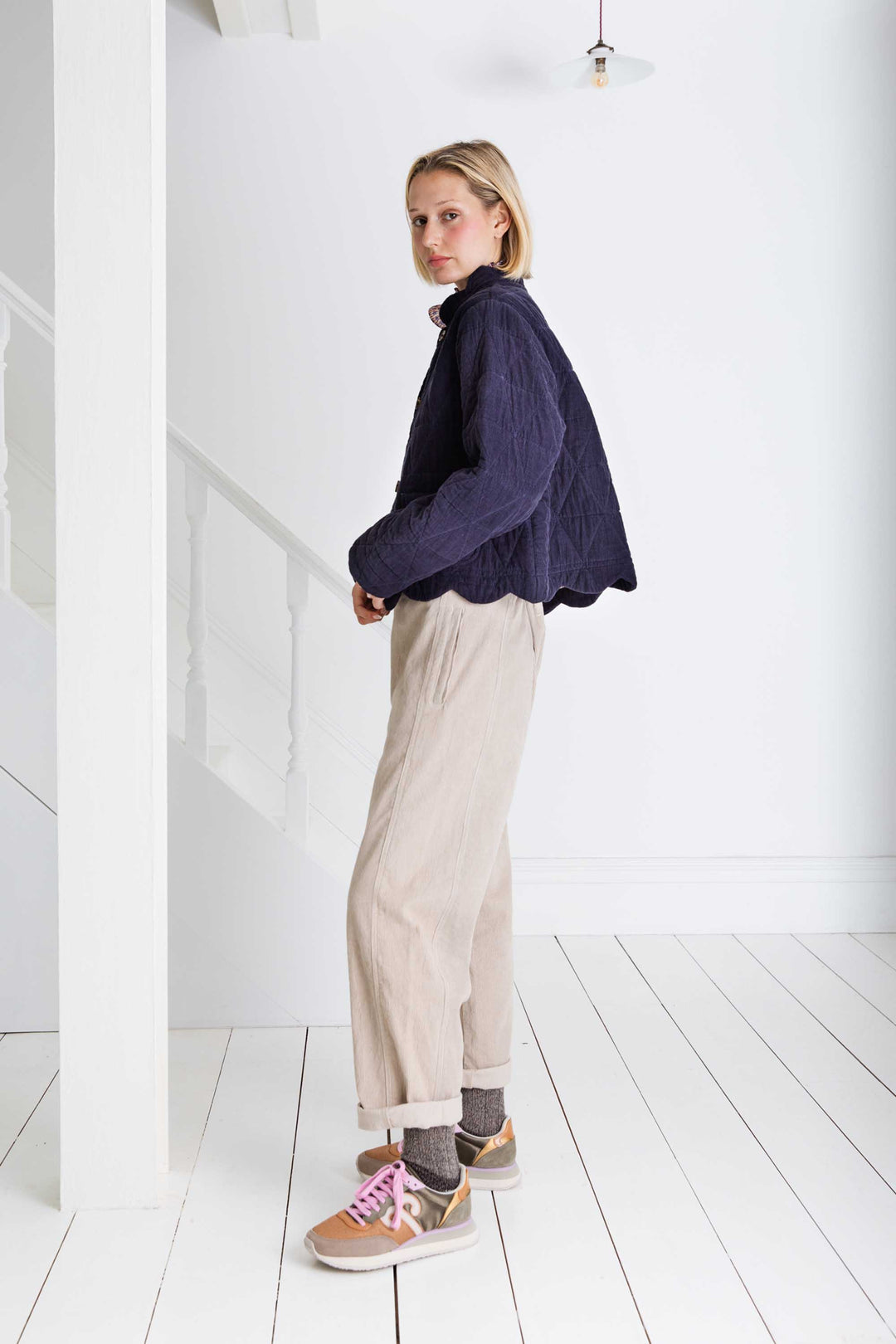 Woman wearing a dark blue knitted sweater and beige pants in a minimal white room.